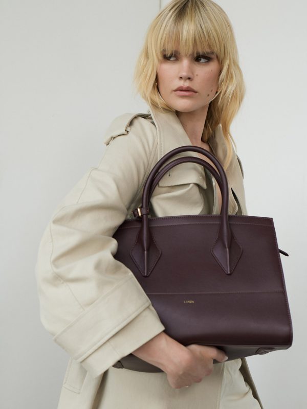 Palto City Bag in Burgunderrot, von weiblichem Modell in Businesskleidung mit Regenmantel in Crème unter dem Arm getragen