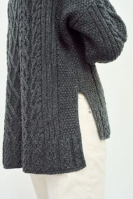 Aran-Pullover in Grau in Kastenform mit Seitenschlitzen. Weibliches Modell mit weissen Hosen trägt Aran Kastenpullover, Seitenansicht von Nahem