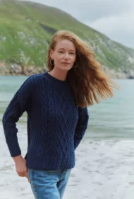 Aran-Pullover in Blau in Kastenform mit Seitenschlitzen. Weibliches Modell mit Jeans und schwarzen Boots trägt Aran Kastenpullover am Strand in Irland, Nahaufnahme 2