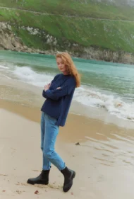 Aran-Pullover in Blau in Kastenform mit Seitenschlitzen. Weibliches Modell mit Jeans und schwarzen Boots trägt Aran Kastenpullover am Strand in Irland