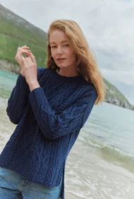 Aran-Pullover in Blau in Kastenform mit Seitenschlitzen. Weibliches Modell mit Jeans und schwarzen Boots trägt Aran Kastenpullover am Strand in Irland, Nahaufnahme